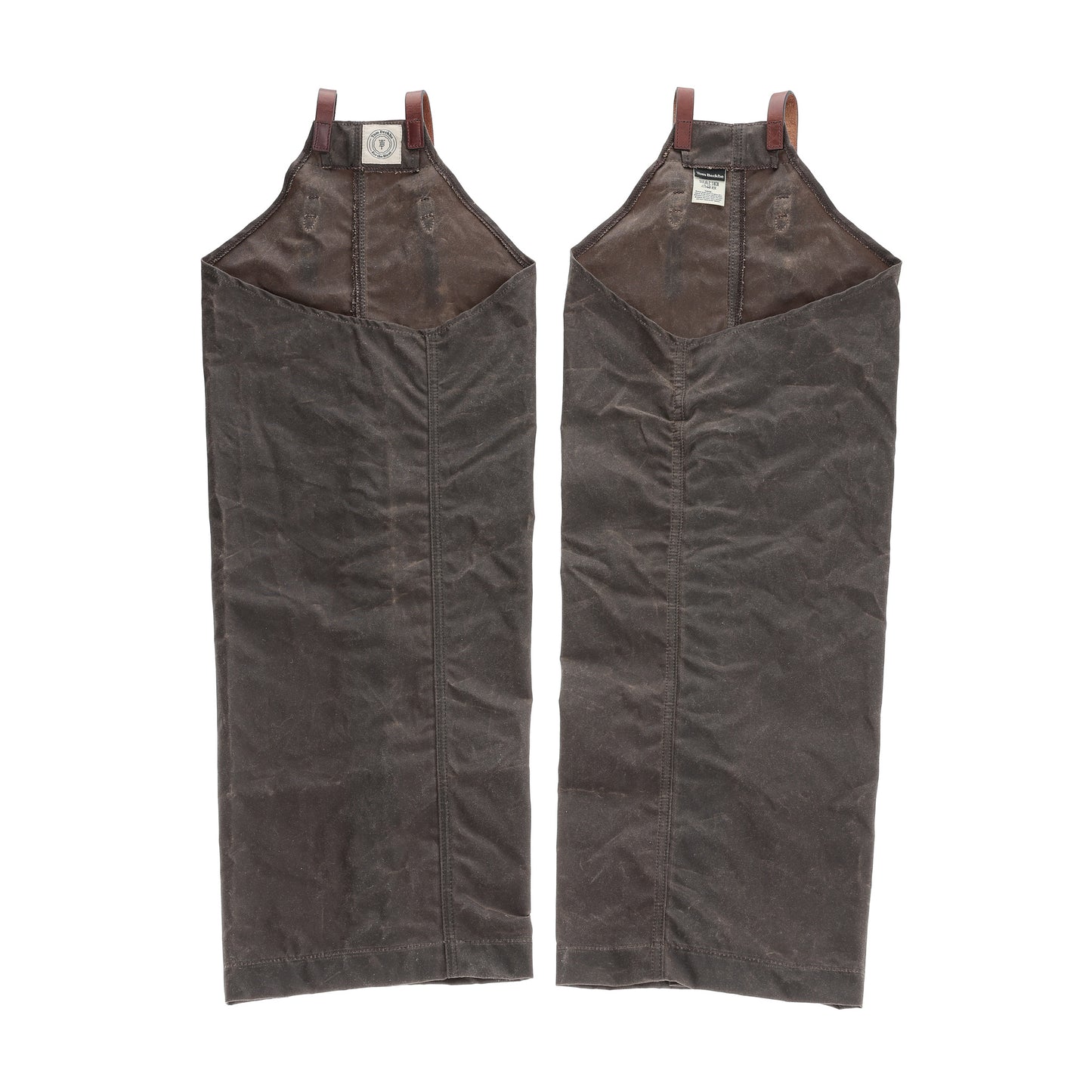 Two brown Tom Beckbe Waxed Canvas Chaps with leather trim and loops lay flat side by side on a white background.