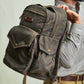 Someone holds the Tom Beckbe DU Canvas Backpack with multiple pockets, brass buttons, and a padded laptop sleeve.