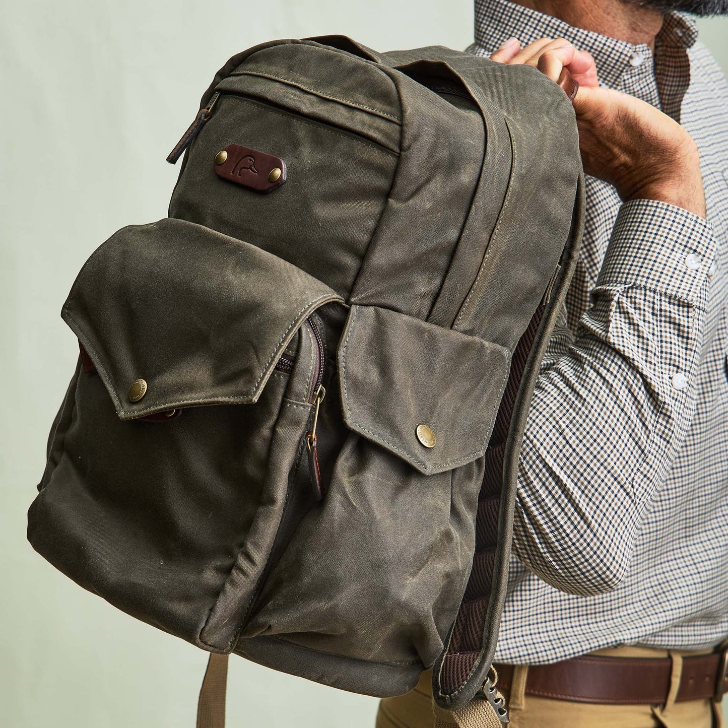 Someone holds the Tom Beckbe DU Canvas Backpack with multiple pockets, brass buttons, and a padded laptop sleeve.