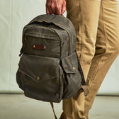 Person holding the Tom Beckbe DU Canvas Backpack with padded laptop sleeve by the top handle, next to beige pants.