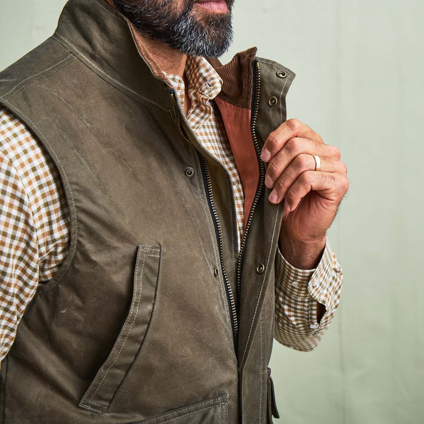 Man wearing a brown plaid shirt and Tom Beckbe DU Kinsman Vest, holding it open to show the waxed cotton construction.