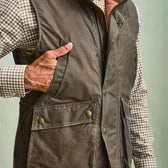 Man wearing a Tom Beckbe DU Kinsman Vest, a brown waxed cotton vest, over a plaid shirt with one hand in the chest pocket.