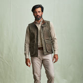 A man stands against a plain background wearing a Tom Beckbe DU Kinsman Vest over a plaid shirt and beige pants.