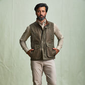 Man stands against a plain background wearing a Tom Beckbe DU Kinsman Vest, checkered shirt, and beige pants.