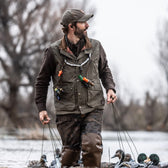 A man in a Tom Beckbe DU Kinsman Vest stands in shallow water with decoy ducks and trees softly rising behind him.