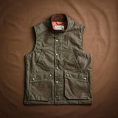 Tom Beckbe DU Kinsman Vest in olive waxed cotton with snap buttons and flap pockets, shown on a brown surface.