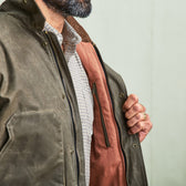 Man opening an olive green jacket to show a zippered interior pocket and checked shirt underneath.