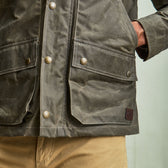Close-up of a person wearing an olive green waxed jacket with snap pockets and tan pants, hand in pocket.