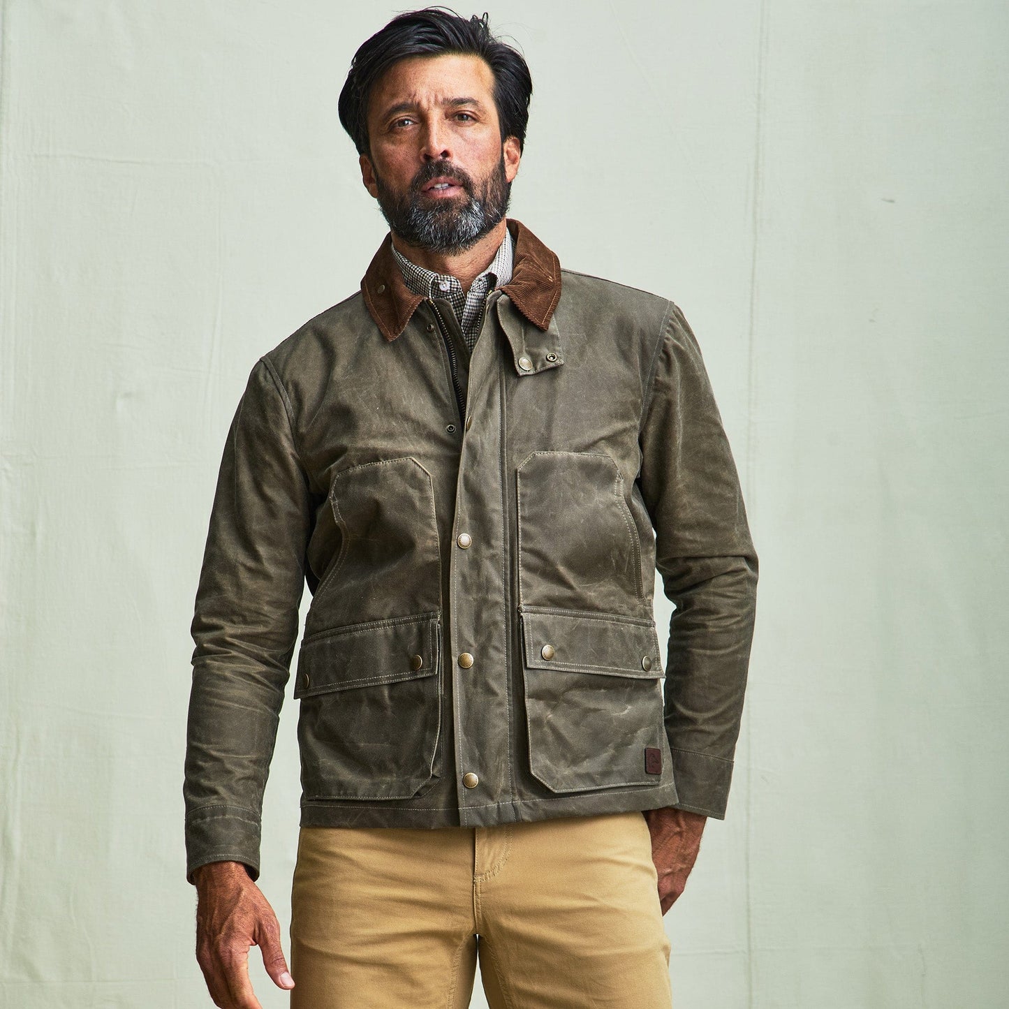 A man wears a Tom Beckbe Steward Jacket with a brown collar, plaid shirt, and tan pants against a light background.