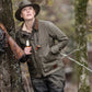 A woman in a Tom Beckbe Womens Steward Jacket leans against a tree with a rifle, gazing upward in the forest.