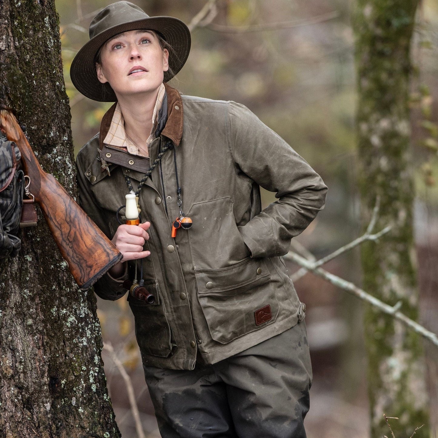 A woman in a Tom Beckbe Womens Steward Jacket leans against a tree with a rifle, gazing upward in the forest.