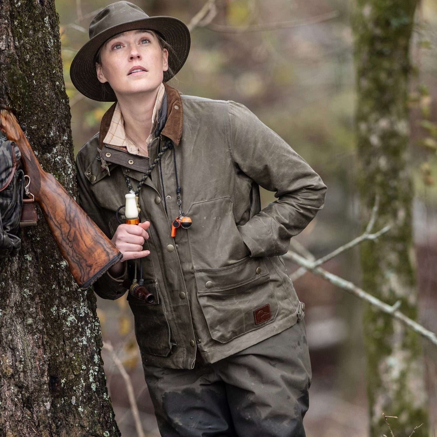A woman in a Tom Beckbe Womens Steward Jacket leans against a tree with a rifle, gazing upward in the forest.