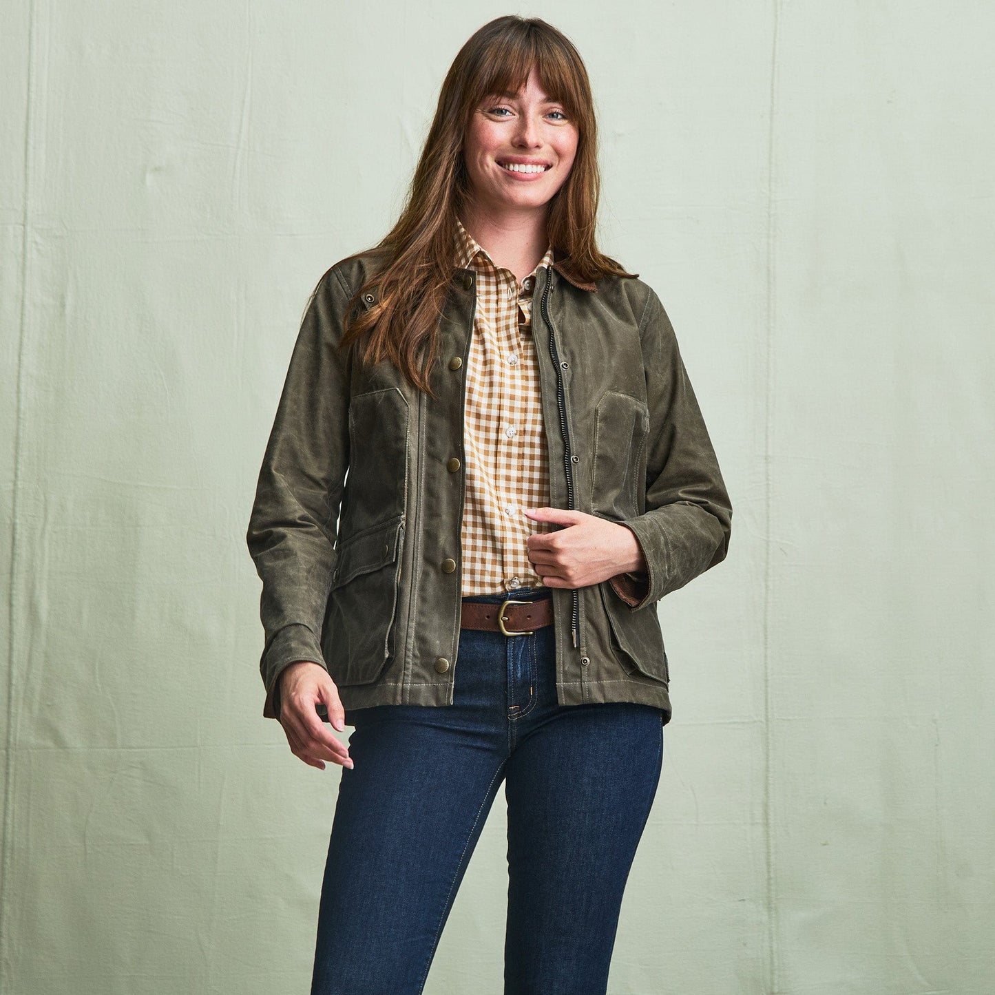 A woman wears the Tom Beckbe Womens Steward Jacket, plaid shirt, and jeans, smiling before a plain light background.