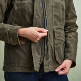 Person zipping up an olive green waxed jacket with front pockets, wearing dark blue jeans.