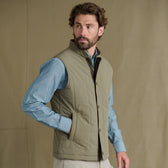Man wearing a light olive quilted vest over a blue shirt stands against a plain beige background, looking to his left.