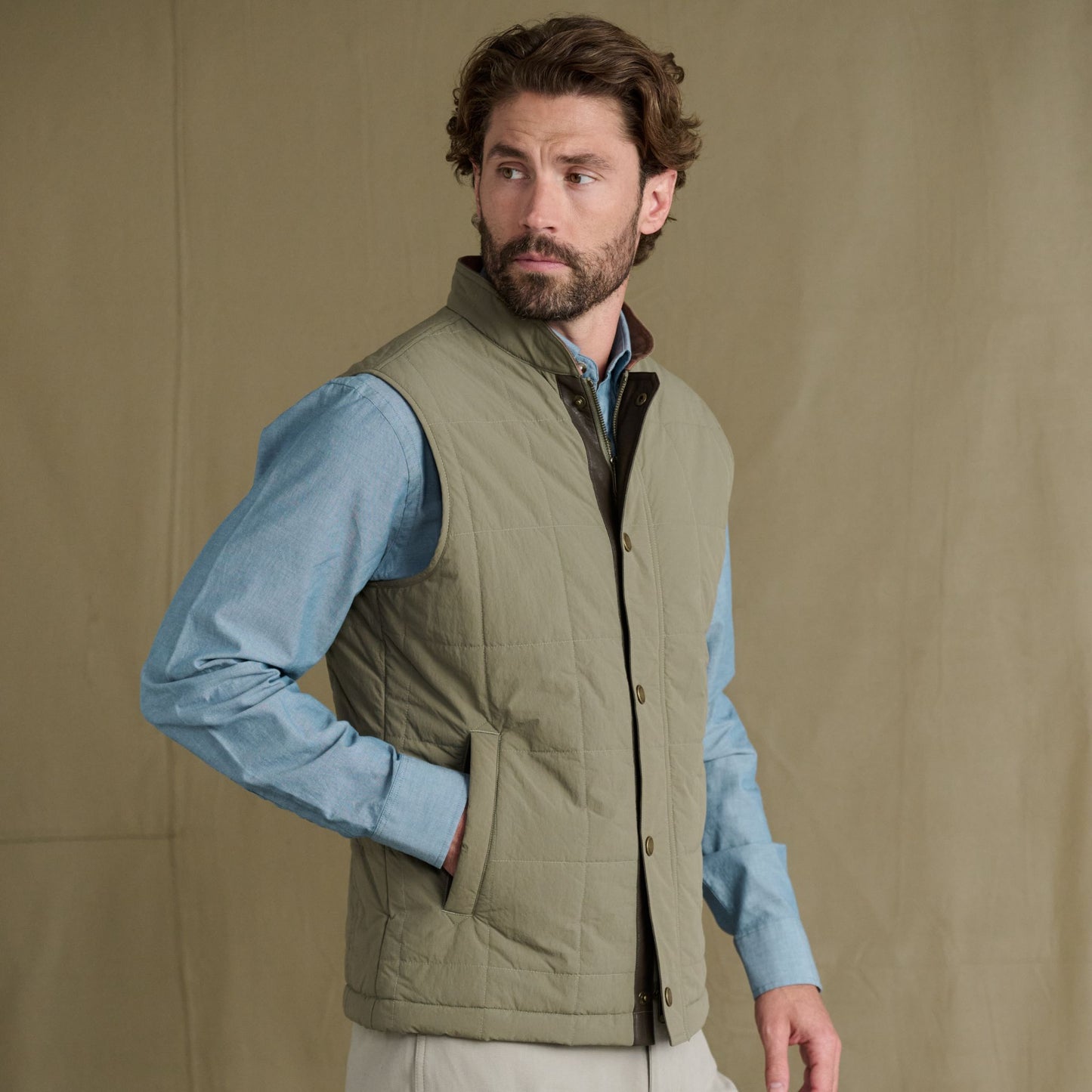 Man wearing a light olive quilted vest over a blue shirt stands against a plain beige background, looking to his left.