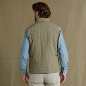 Man facing away, wearing a light green quilted vest over a blue shirt and beige pants, standing against a tan background.