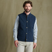 Man wearing a blue quilted vest over a plaid shirt and beige pants stands in front of a plain tan background.