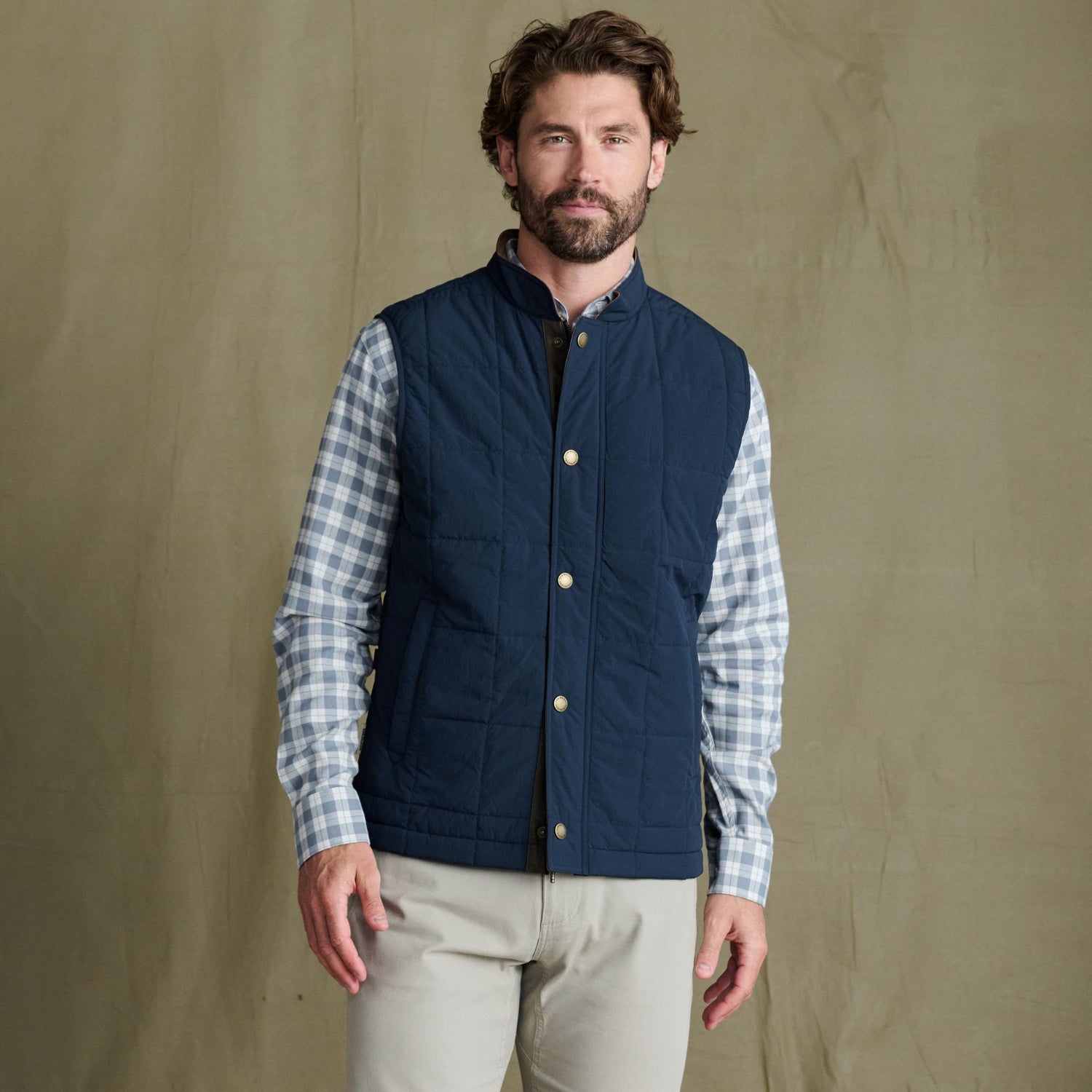 Man wearing a blue quilted vest over a plaid shirt and beige pants stands in front of a plain tan background.