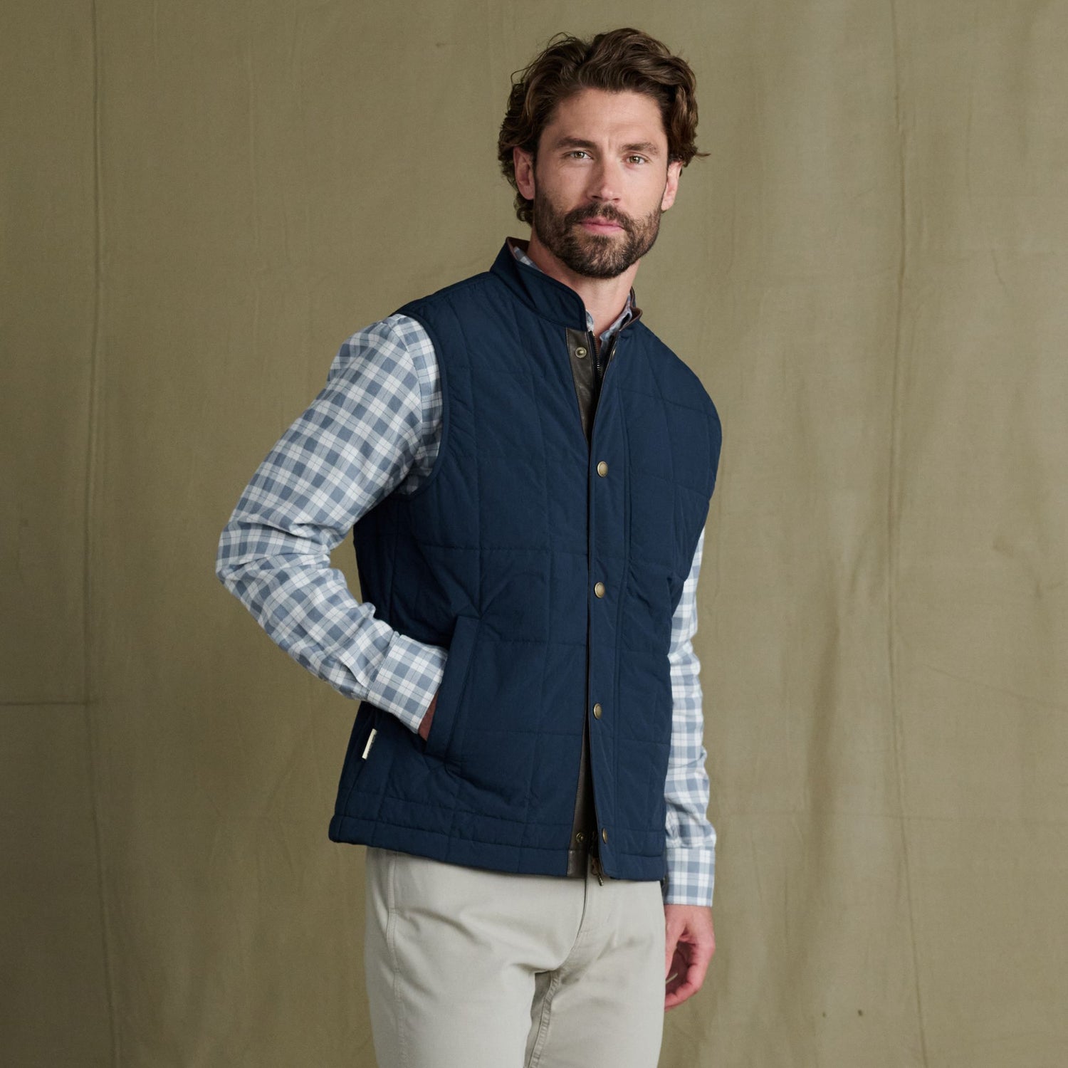 Man wearing a blue quilted vest over a plaid shirt and light pants, standing against a plain beige backdrop.