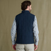 Man facing away, wearing a navy quilted vest over a light plaid shirt and beige pants, standing against a plain background.