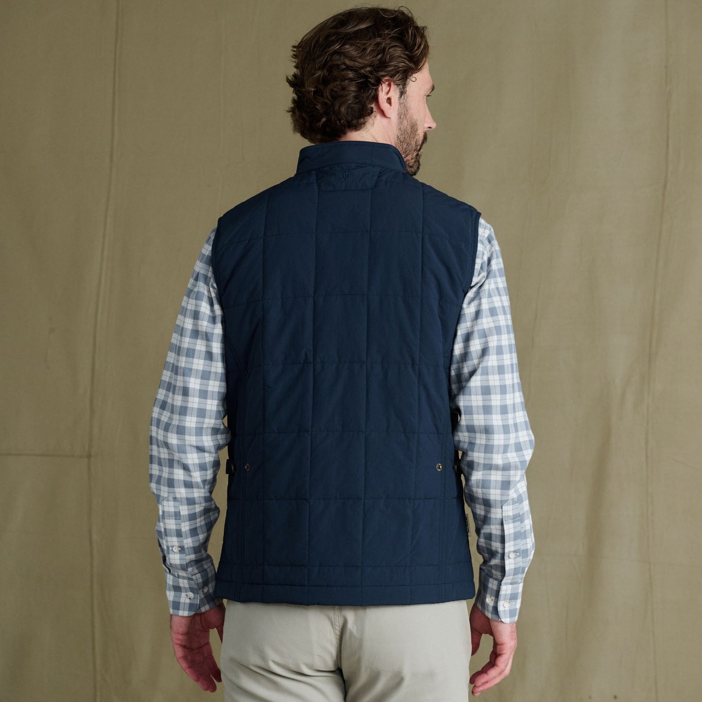 Man facing away, wearing a navy quilted vest over a light plaid shirt and beige pants, standing against a plain background.