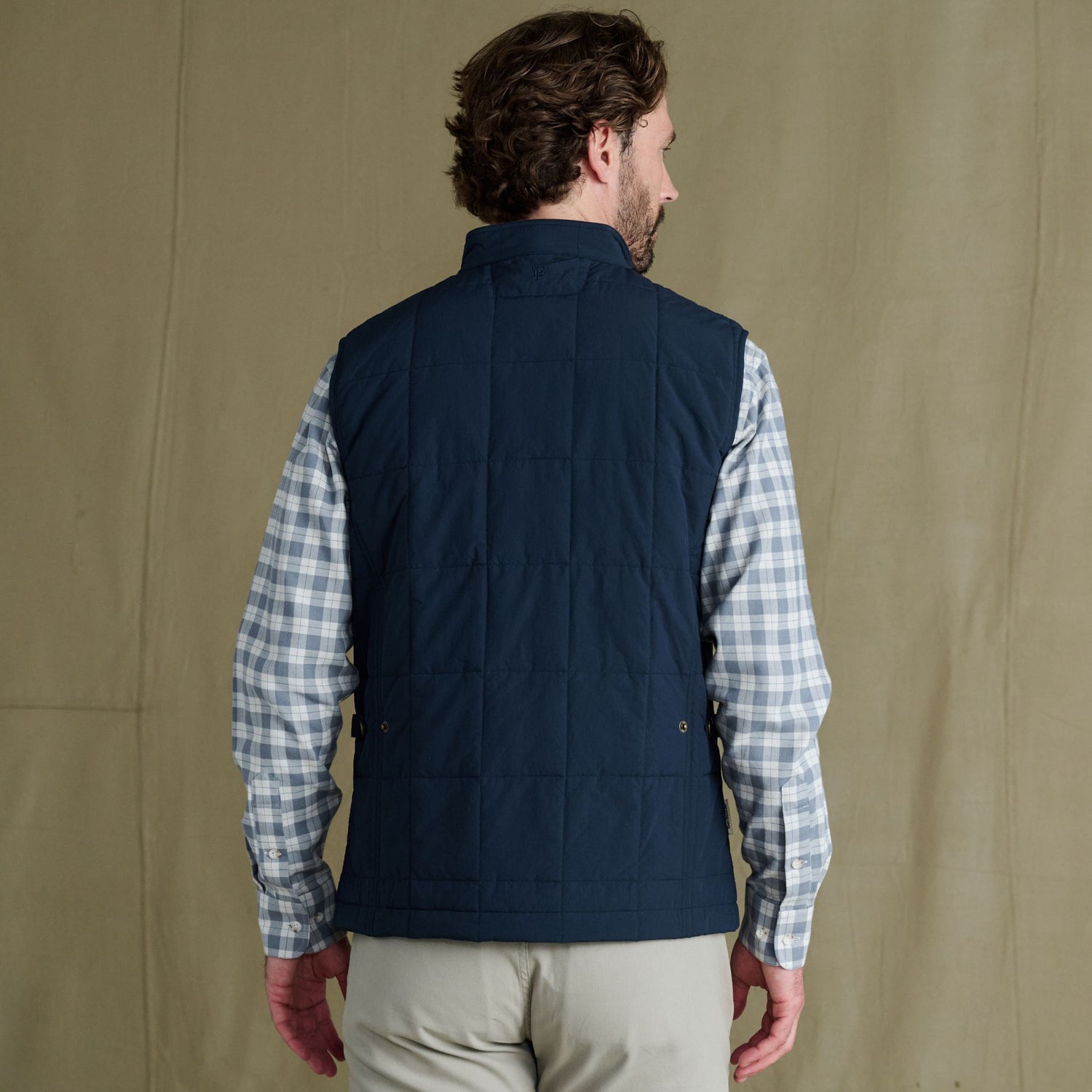 Man facing away, wearing a navy quilted vest over a light plaid shirt and beige pants, standing against a plain background.