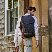 Man with a black backpack opens a wooden door on a shingled building, wearing a plaid shirt and beige pants.