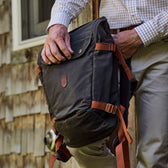 A person in a plaid shirt and beige pants holds a black backpack with brown straps while standing outside near a wooden wall.