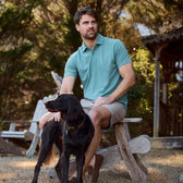 Man in a Coastal Polo Shirt by Tom Beckbe, khaki shorts, sitting outside on a wooden chair with a black dog; trees in the background.