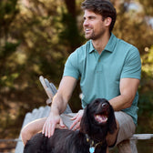 A man in a Tom Beckbe Coastal Polo Shirt on a bench with his black dog, smiling with trees in the backdrop.