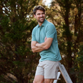 A man in a Tom Beckbe Coastal Polo Shirt and beige shorts leans against a railing with trees in the background.