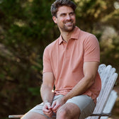 Someone sitting outside on a wooden chair in a Tom Beckbe Coastal Polo Shirt and beige shorts, with greenery around.