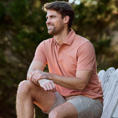 A man sits on a wooden chair, wearing a Tom Beckbe Coastal Polo Shirt and khaki shorts, smiling to his right.