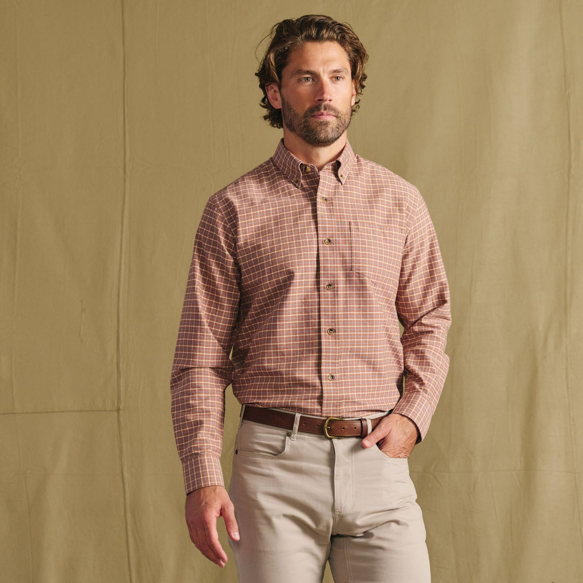 Man standing against a tan backdrop, wearing a red checkered button-up shirt and light-colored pants with a brown belt.