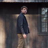 Man in a dark quilted jacket and khaki pants standing outside in front of a wooden building with a window.