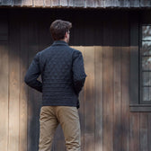 Man in a black quilted jacket and khaki pants stands facing a wooden building with his back to the camera.