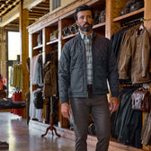 A man in a quilted jacket and slacks walks through a clothing store with wooden shelves and hanging jackets.