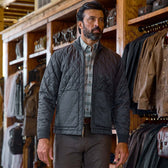 Man wearing a quilted gray jacket and plaid shirt stands in a clothing store with racks of jackets in the background.