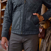 A person wearing a quilted black jacket and gray pants stands indoors with one hand in a jacket pocket.