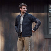 Man wearing a quilted black jacket, plaid shirt, and khaki pants stands in front of a dark wooden building.