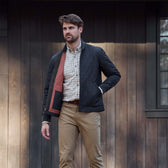 Man wearing a quilted black jacket, checked shirt, and khaki pants stands in front of a dark wooden wall.