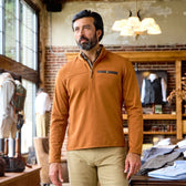 A man wearing a Tom Beckbe Tucker Fleece Quarter Zip and khaki pants stands in a store with exposed brick walls.