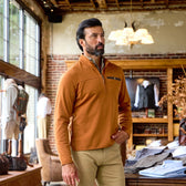 A man wears the Tom Beckbe Tucker Fleece Quarter Zip and khaki pants in a store with casual outdoor apparel on display.