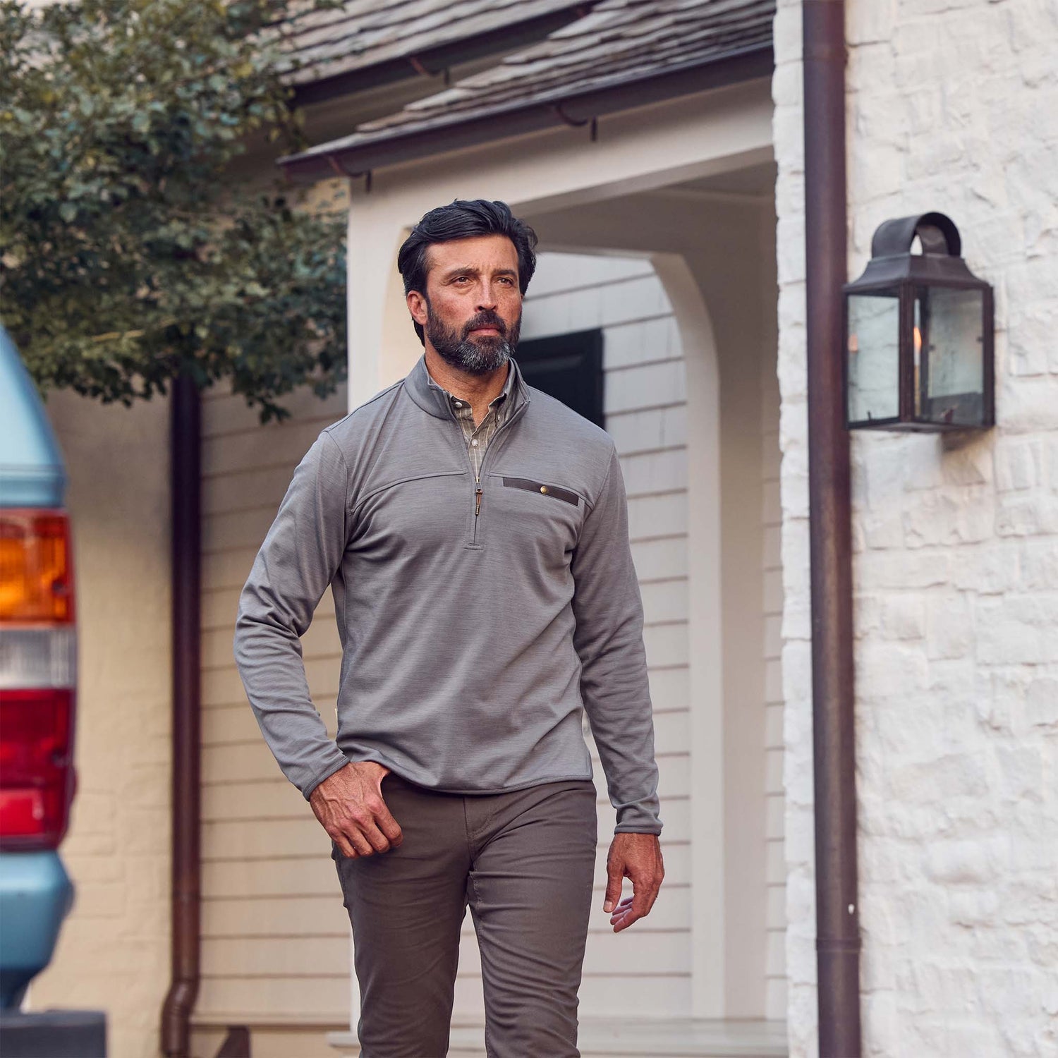 A bearded man walks outside near a white house, wearing Tom Beckbes Tucker Fleece Quarter Zip and brown pants.
