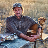 Man in plaid shirt and Tom Beckbe Waxed Cotton 6-Panel Baseball Hat sits outside with empty plates; dog stands nearby in field.