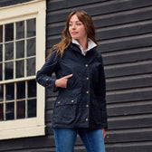 Woman in a navy jacket with pockets and jeans stands by a dark wooden building, looking to the side.