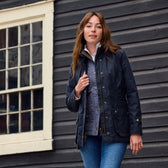 A woman in a Tom Beckbe Womens Blakeley Jacket and blue jeans stands by a black building with a white-framed window.