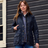 A woman in a navy Tom Beckbe Womens Blakeley Jacket stands against a dark wooden wall, gazing confidently at the camera.
