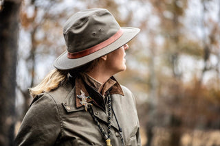 Person in an olive green jacket and hat looks to the side outdoors, with blurred trees in the background.